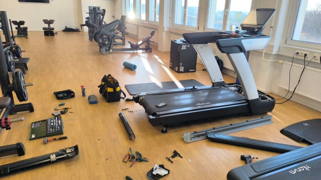 Fitnessruimte met loopband en gereedschap op de vloer tijdens reparatie en onderhoud van fitnessapparatuur.