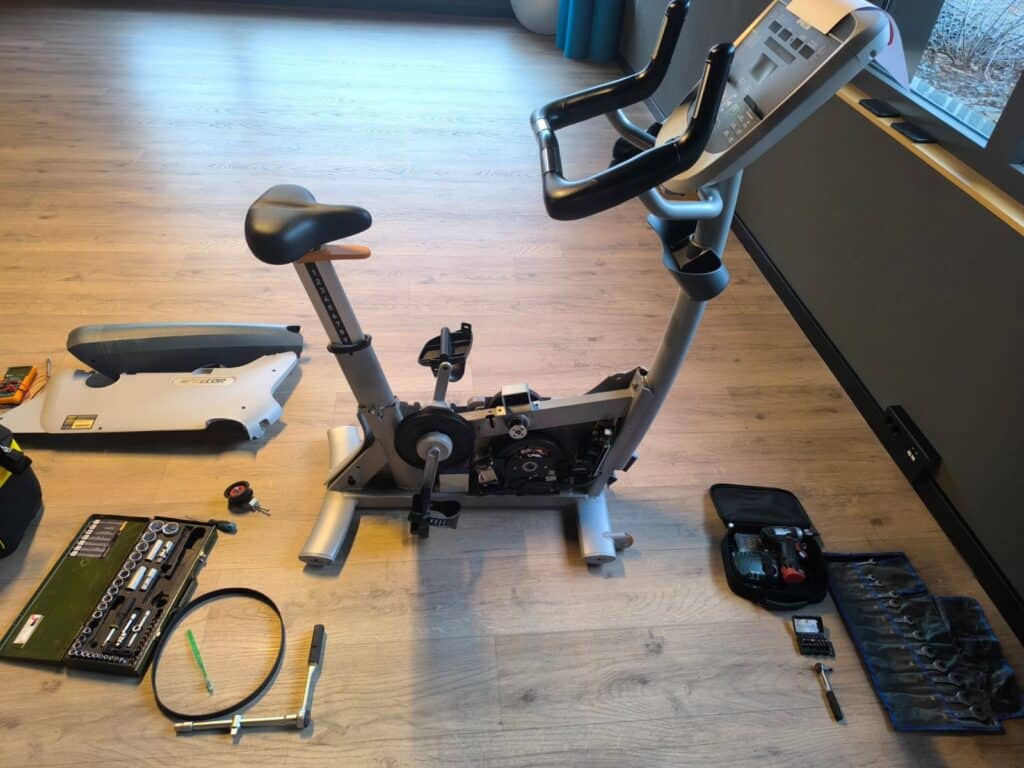Professionele fitnessfiets met open behuizing tijdens reparatie, met gereedschap en onderdelen op de vloer.